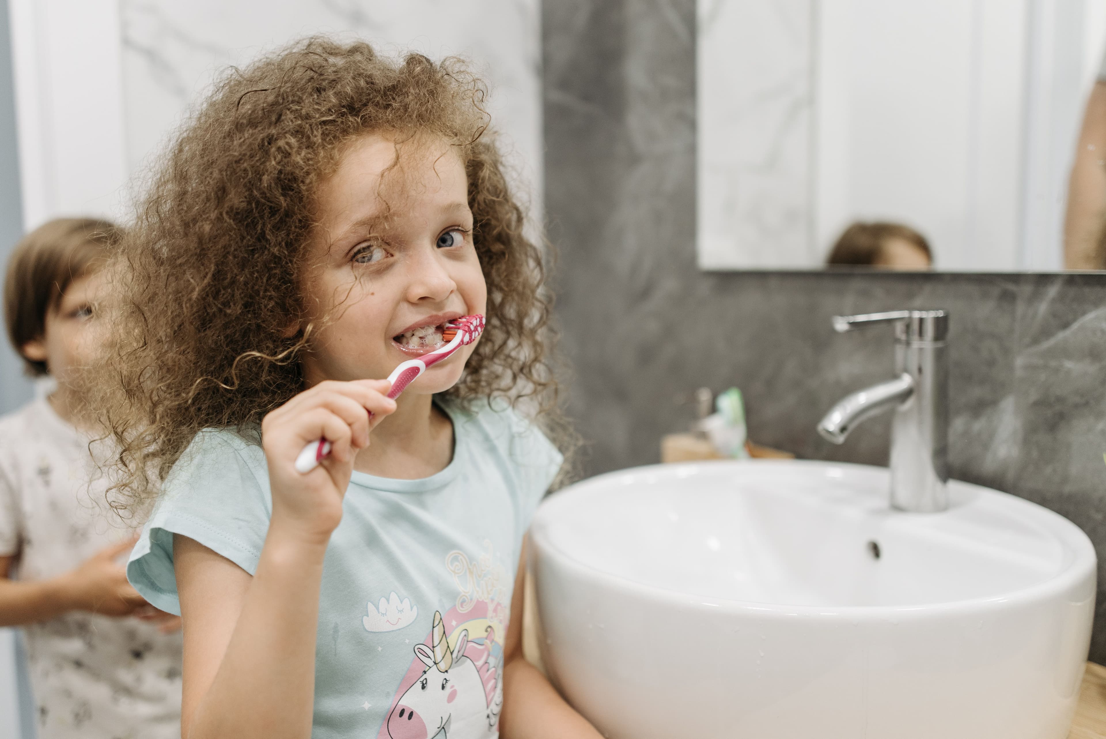 A young girl smiling while brushing her teeth at a bathroom sink