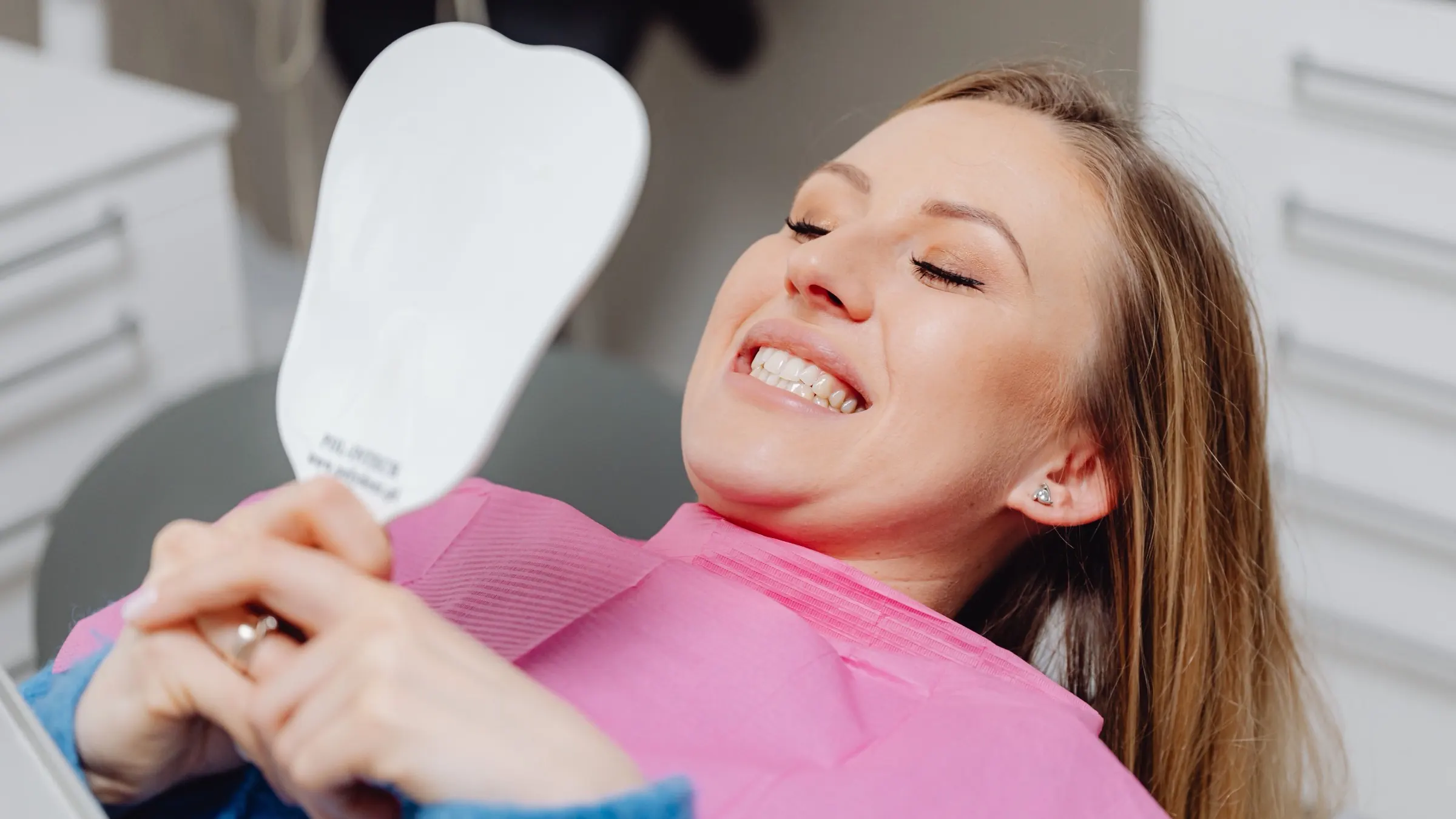 A woman reclined in the dental chair, smiling at her teeth in a hand mirror.